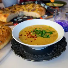 En skål med varm shorbet adas, toppad med persilja och chiliflakes, bredvid ett solrosbröd och focaccia.