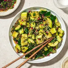 En skål med krossad gurksallad, toppad med sesamfrön och färska örter, redo att avnjutas med ätpinnar.