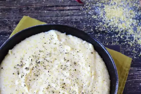Krämig polenta i en gjutjärnspanna, strödd med peppar. Majsgryn syns spridda på den mörka träytan.