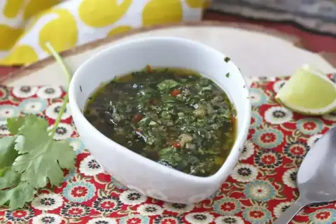 En skål med grön chimichurri, en syrlig limeklyfta och en sked på ett färgglatt tyg.