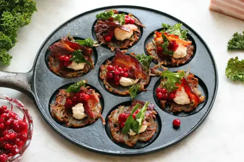 Små, krispiga rårakor med bacon, gräddfil och rårörda lingon i en gjutjärnspanna. Serveras med färska lingon och grönkål.