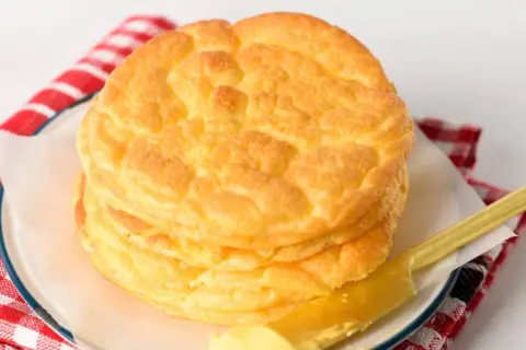 En hög med fluffigt cloud bread på en tallrik, bredvid en rödvitrutig duk och en smörkniv med smör.