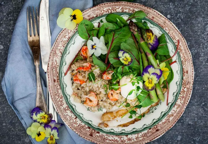 A plate of creamy risotto with shrimp, asparagus, greens, and edible flowers, served with a fork and knife.