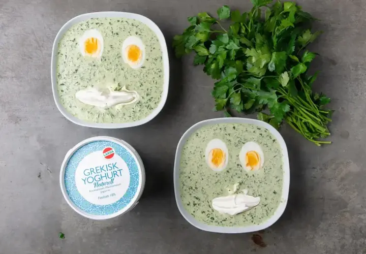Två skålar med spenatsoppa som ser ut som ansikten, med ägg som ögon och gräddfil som mun. Bredvid står en burk grekisk yoghurt och en grön kvist persilja. Ett roligt spenatsoppa recept!
