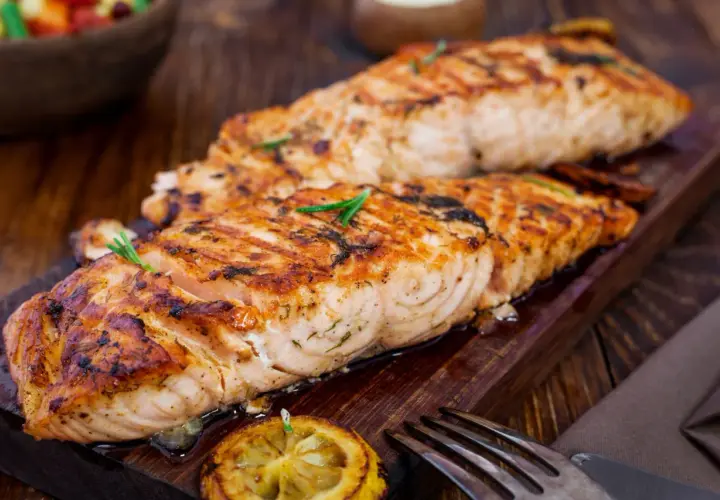 Två saftiga filéer av grillad fisk, perfekt kryddad med örter, serveras på en träbricka med citron och en gaffel.