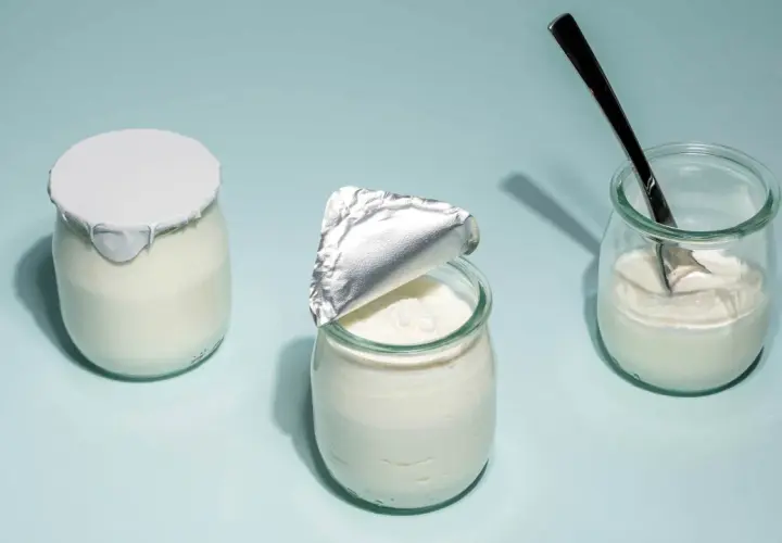 Tre burkar med hemmagjord yoghurt på ljusblå bakgrund. En är helt försluten, en annan är delvis öppnad och en tredje har en sked i sig.