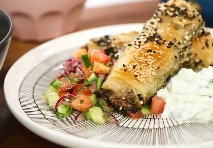 Krispig spenat och fetaostpaj toppad med sesamfrön, serverad med en fräsch tomatsallad och tzatziki.