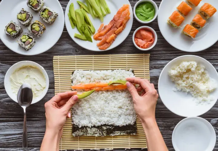 Händer lägger lax och avokado på sushiris för ett sushirecept. Ingredienser som gurka, wasabi och färdig sushi syns också.