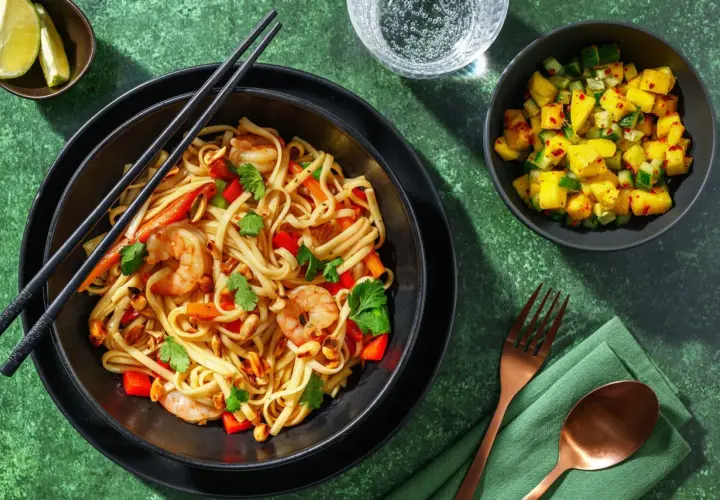 En skål med pad thai med räkor, nudlar, paprika och jordnötter. Serveras med en mangosallad och limeklyftor.
