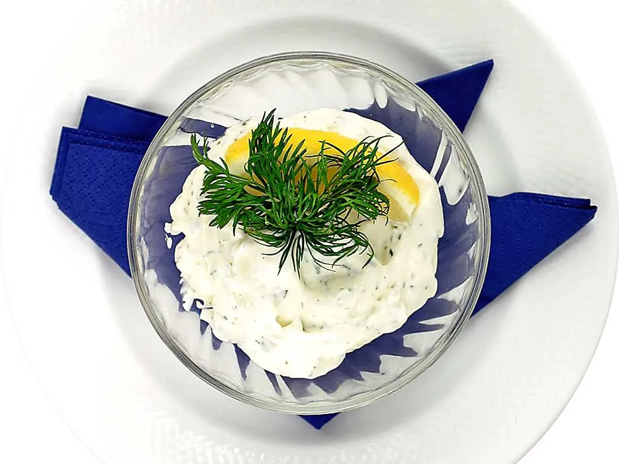 Krämig dillmajonnäs i en skål, garnerad med dill och citron. Serveras på en vit tallrik med en blå servett.