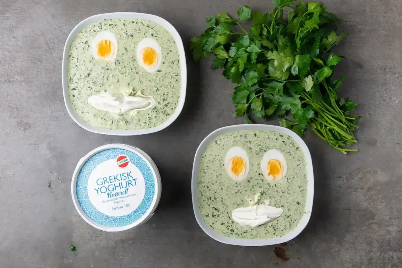 Två skålar med spenatsoppa som ser ut som ansikten, med ägg som ögon och gräddfil som mun. Bredvid står en burk grekisk yoghurt och en grön kvist persilja. Ett roligt spenatsoppa recept!