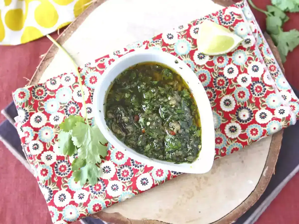 En skål med grön chimichurri, garnerad med en limeklyfta och korianderblad, på en vackert mönstrad duk.